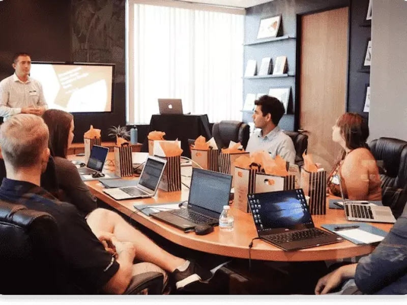 Foto de uma sala de reunião moderna com profissionais sentados em volta de uma mesa, trabalhando em seus notebooks e discutindo estratégias de investimento. Colaboração e tecnologia